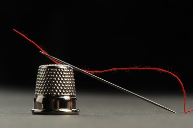 Sewing needle with red thread and thimble on grey table against black background, closeup