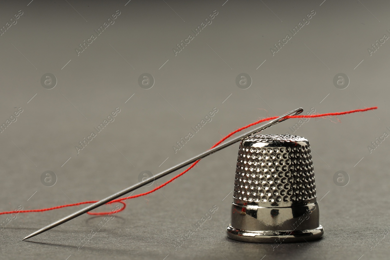 Photo of Sewing needle with red thread and thimble on grey background, closeup. Space for text