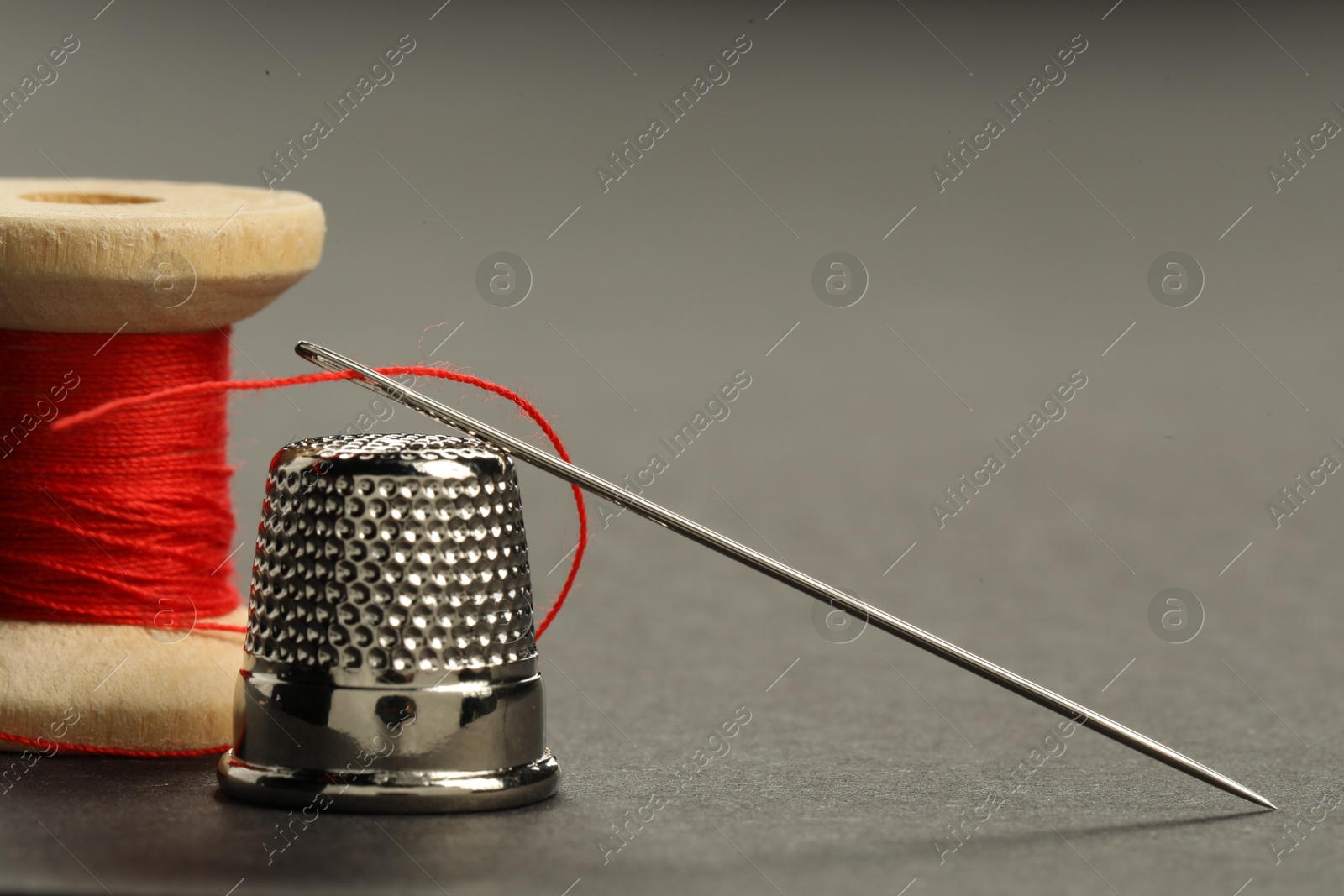 Photo of Sewing needle with red thread, spool and thimble on grey background, closeup. Space for text