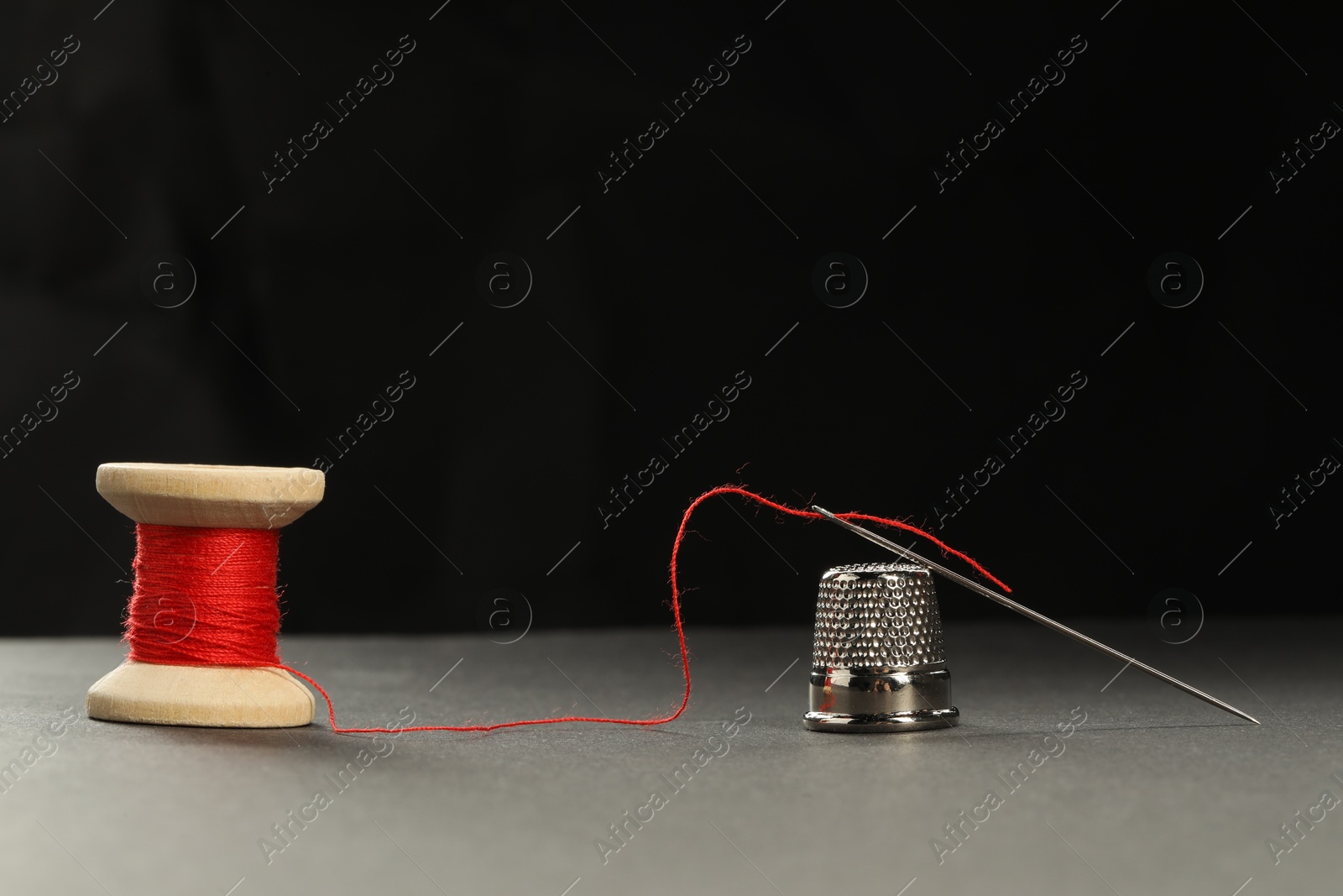 Photo of Sewing needle with red thread and thimble on grey table against black background