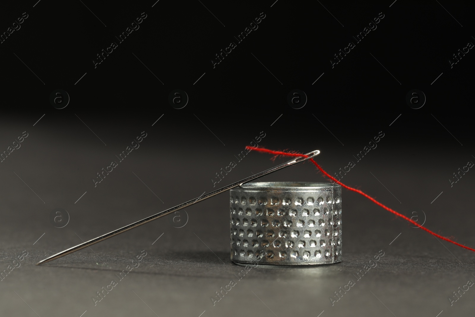 Photo of Sewing needle with red thread and thimble ring on grey table against black background, closeup