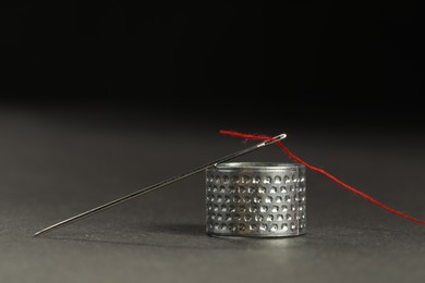 Photo of Sewing needle with red thread and thimble ring on grey table against black background, closeup