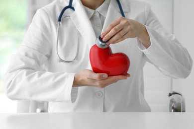 Doctor with stethoscope and red heart at table in clinic, closeup