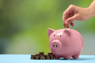 Photo of Woman putting coin into pink piggy bank at light blue table against blurred background, closeup. Space for text