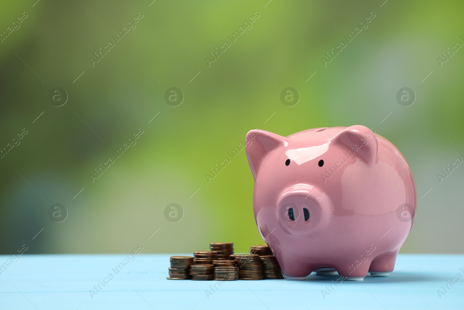 Photo of Pink piggy bank and stacks of coins on light blue table against blurred background, space for text