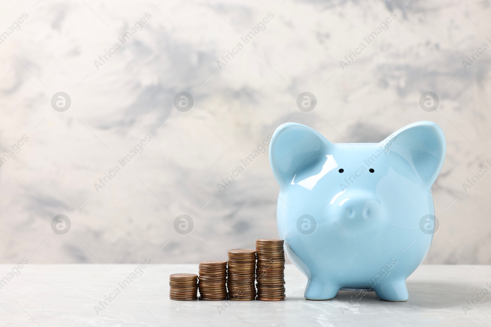 Photo of Blue piggy bank and stacks of coins on light marble table, space for text