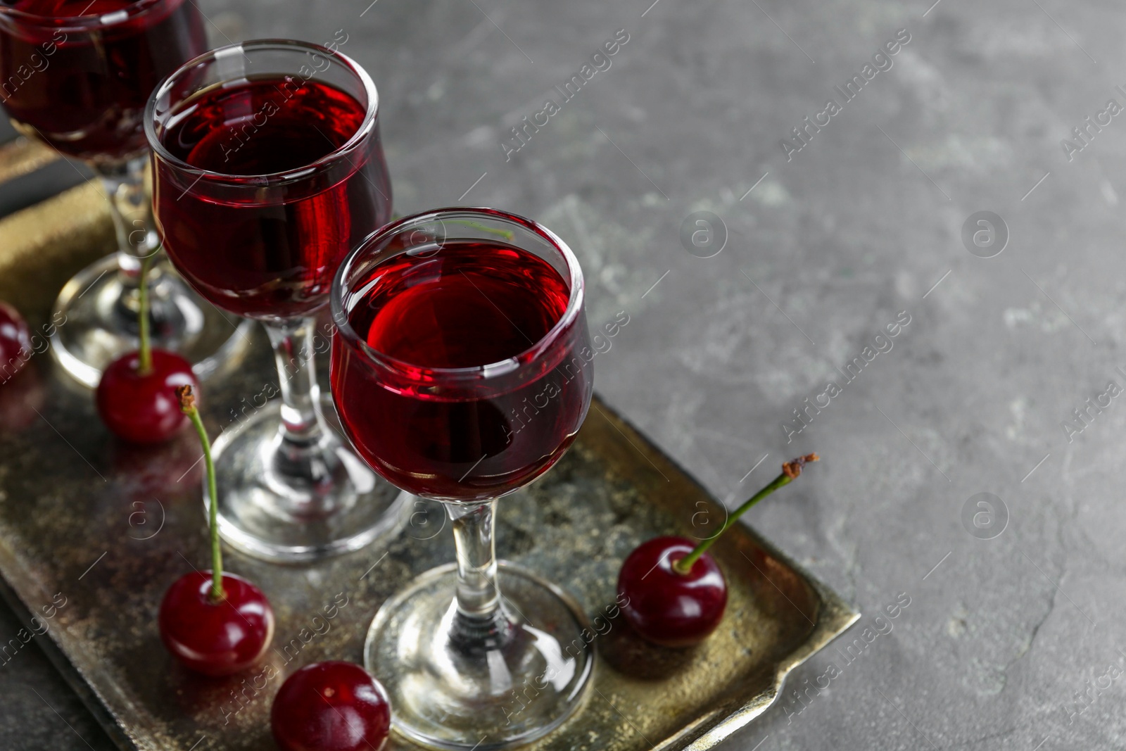 Photo of Delicious cherry liqueur in glasses and fresh berries on grey table, space for text