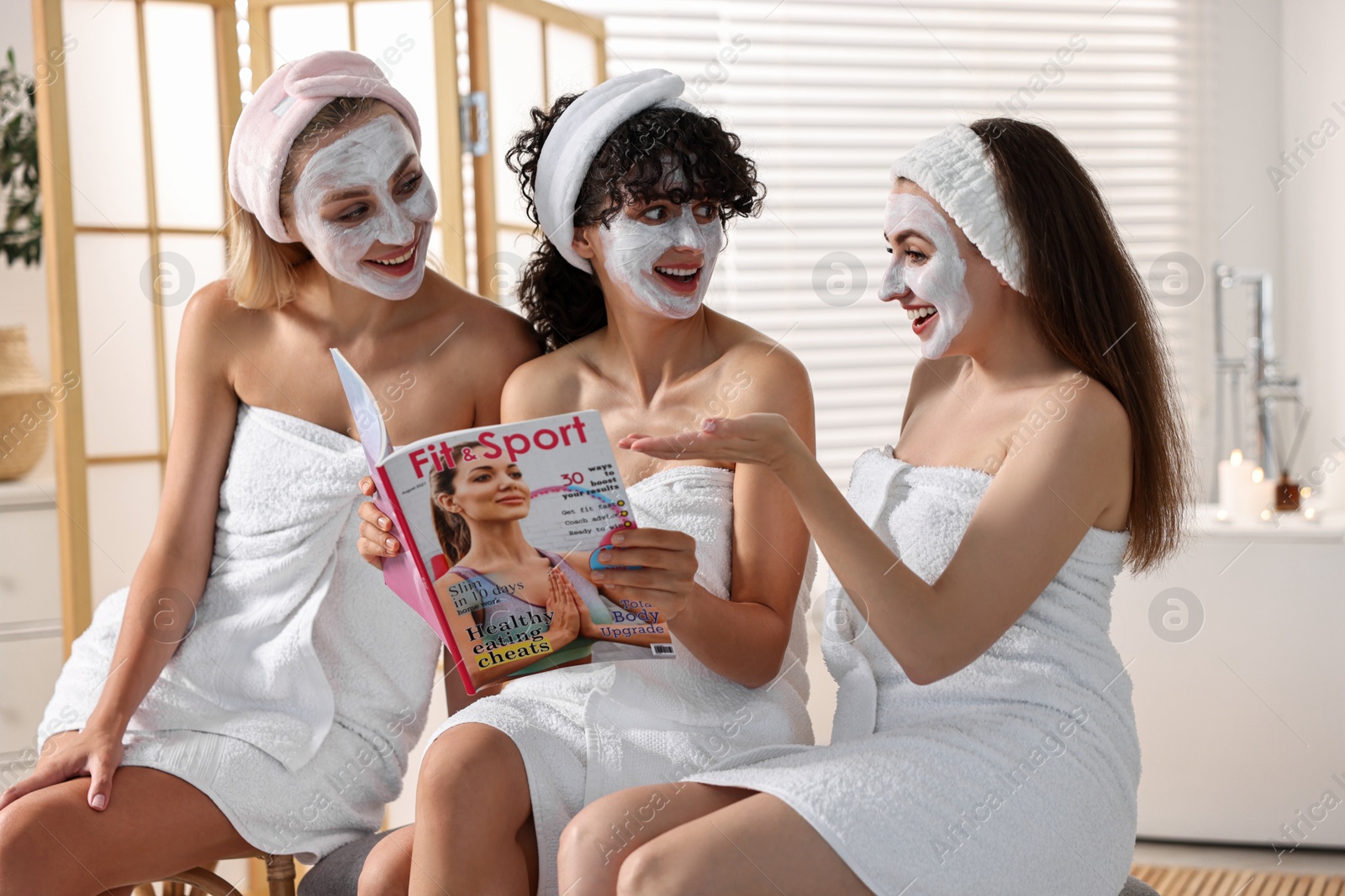 Photo of Happy friends with facial masks reading magazine in bathroom. Spa party