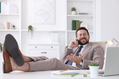 Smiling businessman talking by smartphone and holding legs on table in office