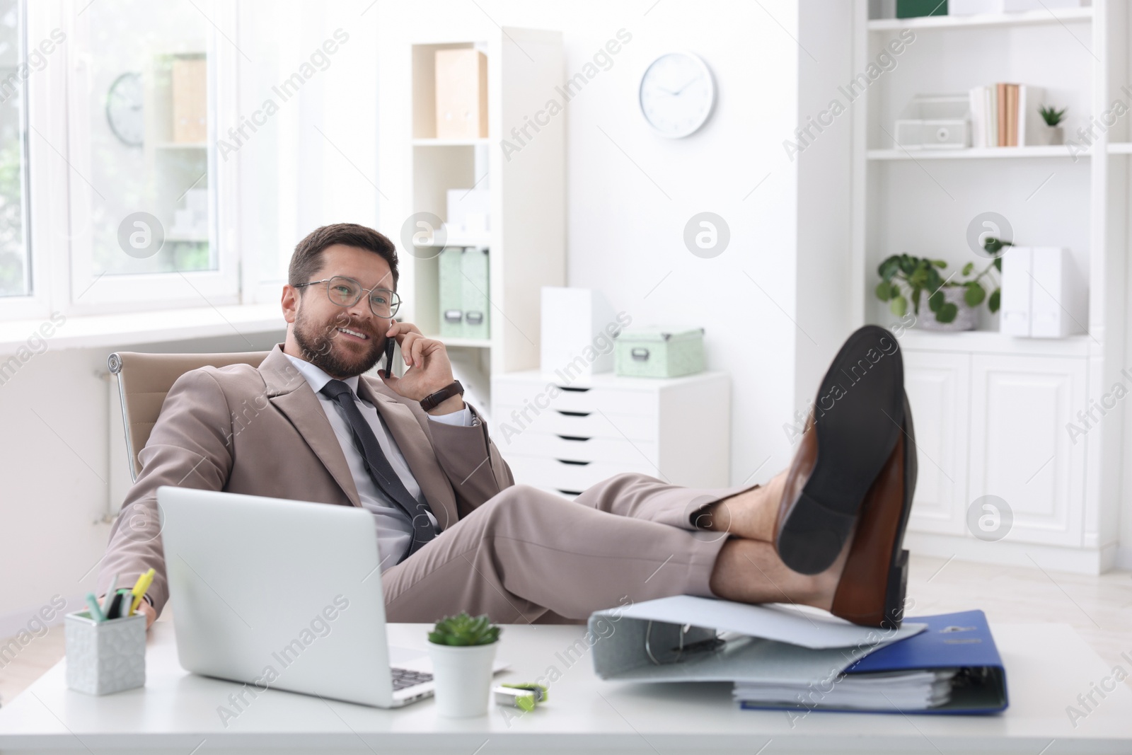 Photo of Smiling businessman talking by smartphone and holding legs on table in office