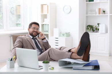 Smiling businessman talking by smartphone and holding legs on table in office