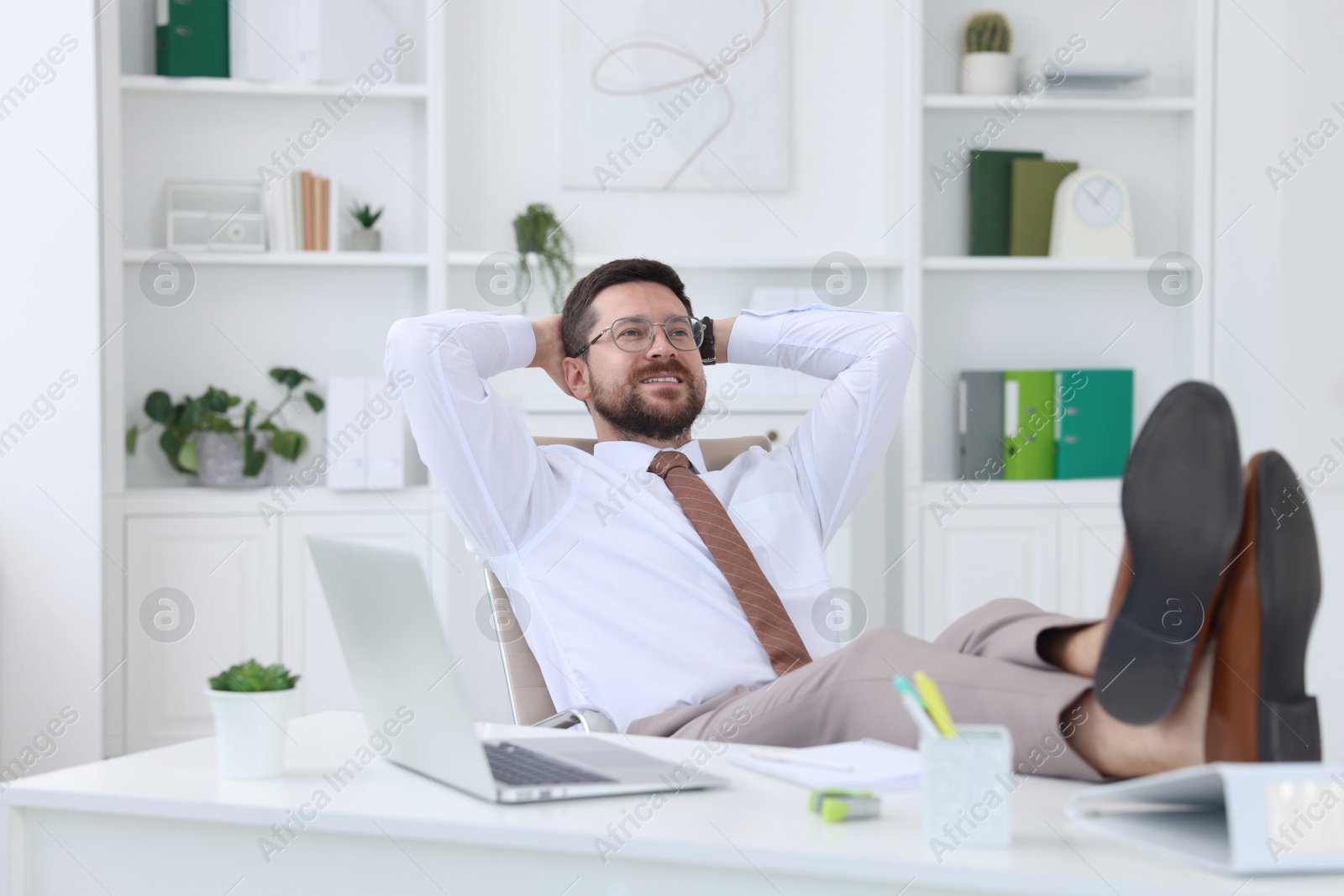 Photo of Smiling businessman with hands behind his head holding legs on table in office. Break time