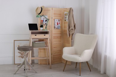 Photo of Folding screen, table, laptop, lamp and armchair indoors. Interior design