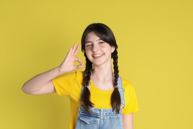 Portrait of smiling teenage girl showing ok gesture on yellow background