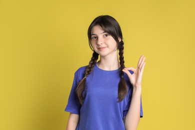 Portrait of cute teenage girl showing ok gesture on yellow background