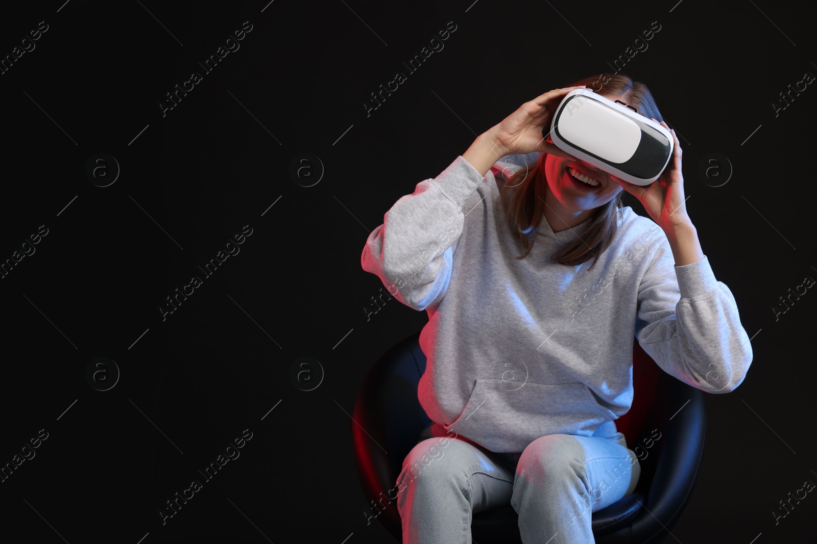 Photo of Happy woman with virtual reality headset sitting on chair in neon lights against black background, space for text