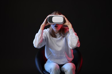 Emotional woman with virtual reality headset sitting on chair in neon lights against black background