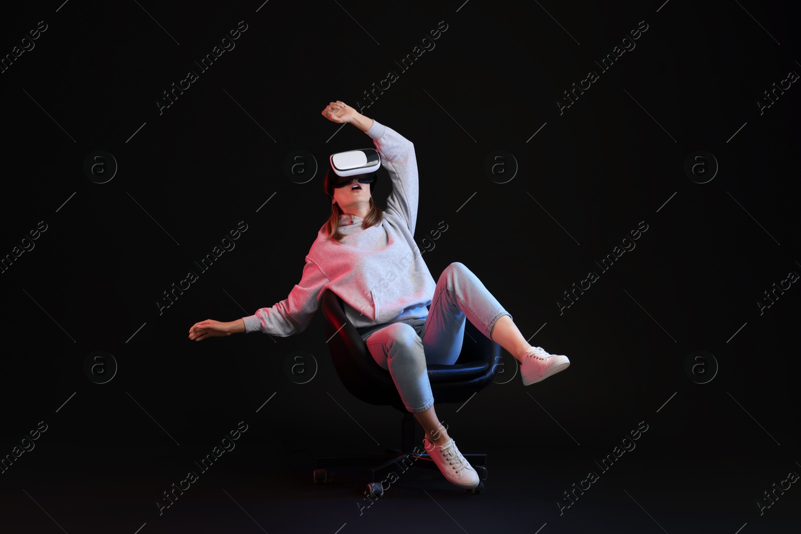 Photo of Emotional woman with virtual reality headset sitting on chair in neon lights against black background