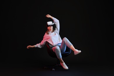 Emotional woman with virtual reality headset sitting on chair in neon lights against black background