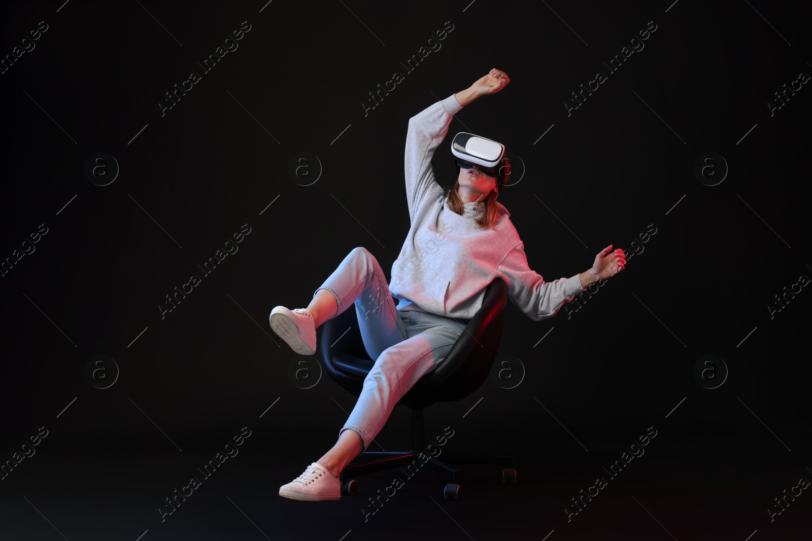 Photo of Emotional woman with virtual reality headset sitting on chair in neon lights against black background