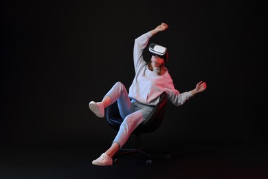 Emotional woman with virtual reality headset sitting on chair in neon lights against black background