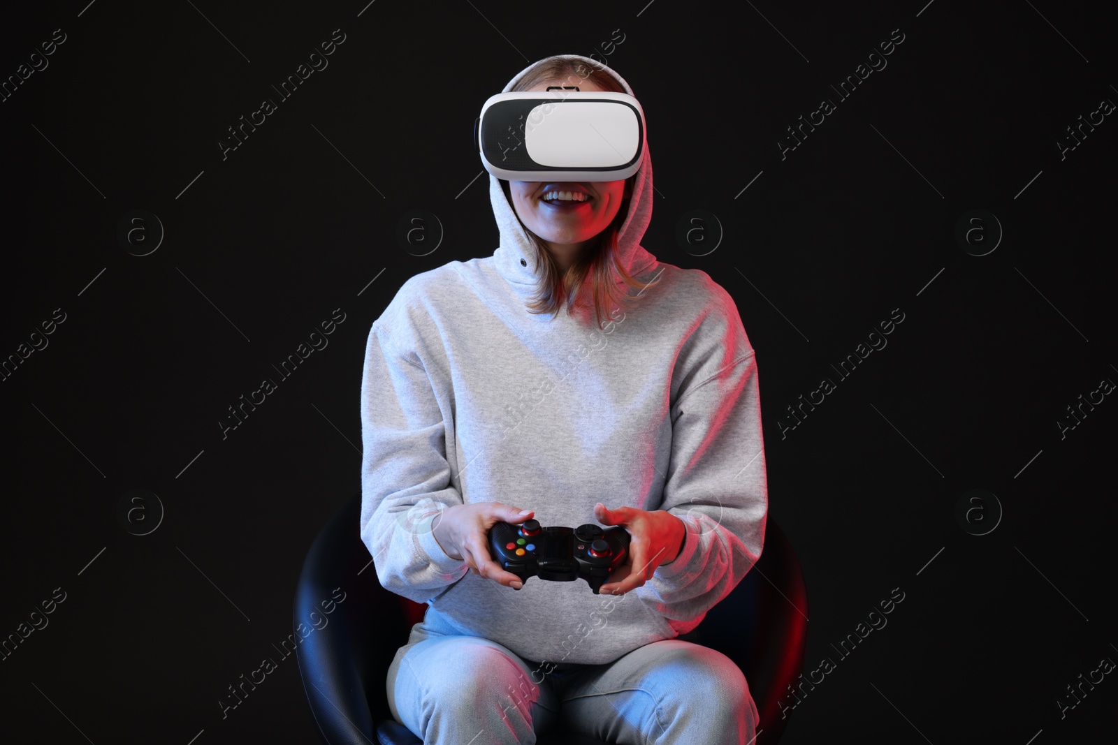 Photo of Happy woman with virtual reality headset and controller sitting on chair in neon lights against black background