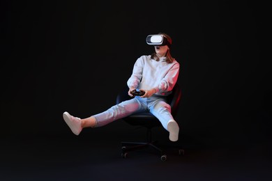 Photo of Emotional woman with virtual reality headset and controller sitting on chair in neon lights against black background