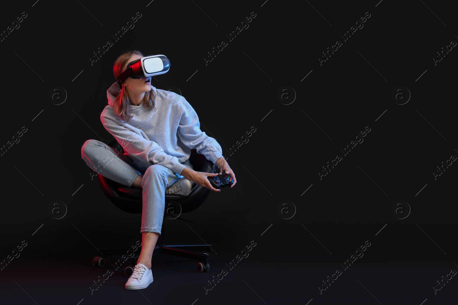 Photo of Emotional woman with virtual reality headset and controller sitting on chair in neon lights against black background, space for text