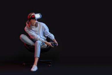 Emotional woman with virtual reality headset and controller sitting on chair in neon lights against black background, space for text