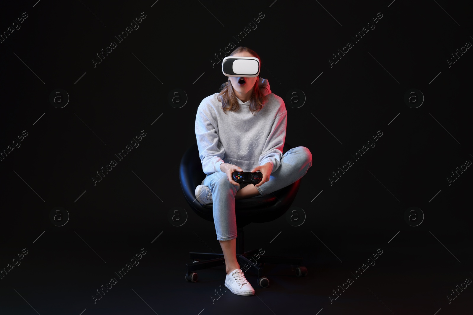 Photo of Emotional woman with virtual reality headset and controller sitting on chair in neon lights against black background