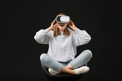 Photo of Happy woman with virtual reality headset sitting on chair against black background