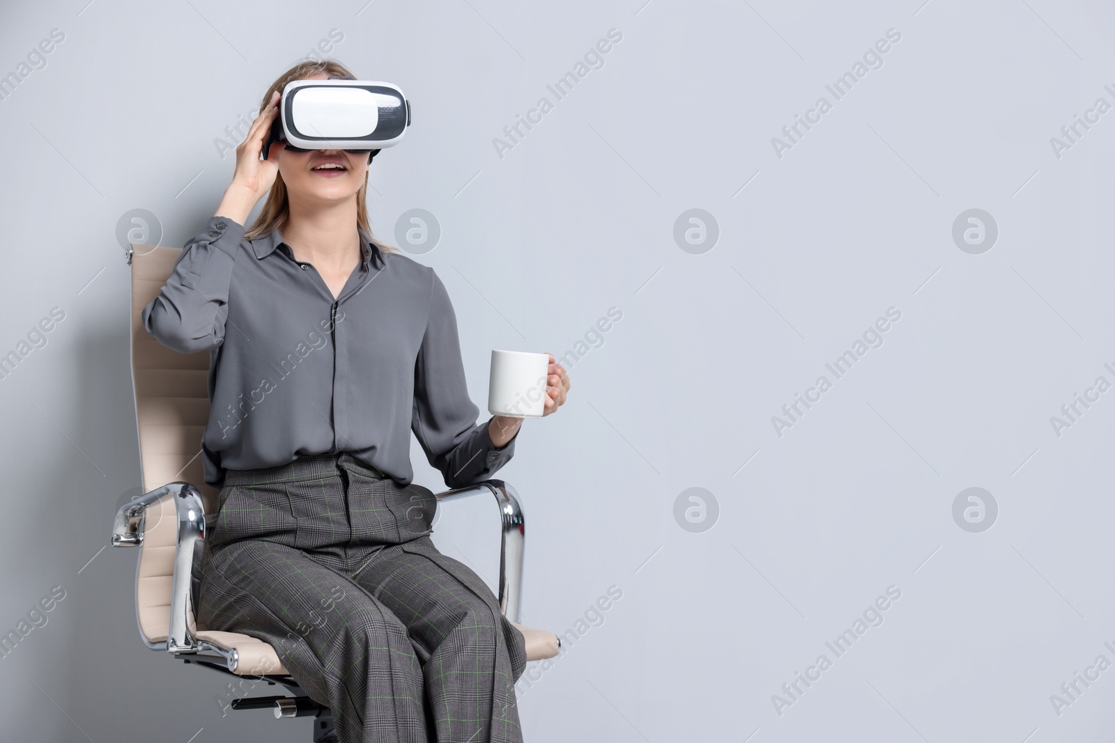 Photo of Happy woman with virtual reality headset and cup sitting on chair against gray background, space for text