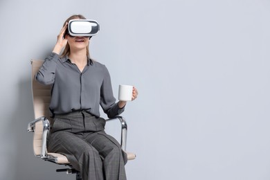 Happy woman with virtual reality headset and cup sitting on chair against gray background, space for text