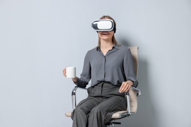 Photo of Happy woman with virtual reality headset and cup sitting on chair against gray background, space for text