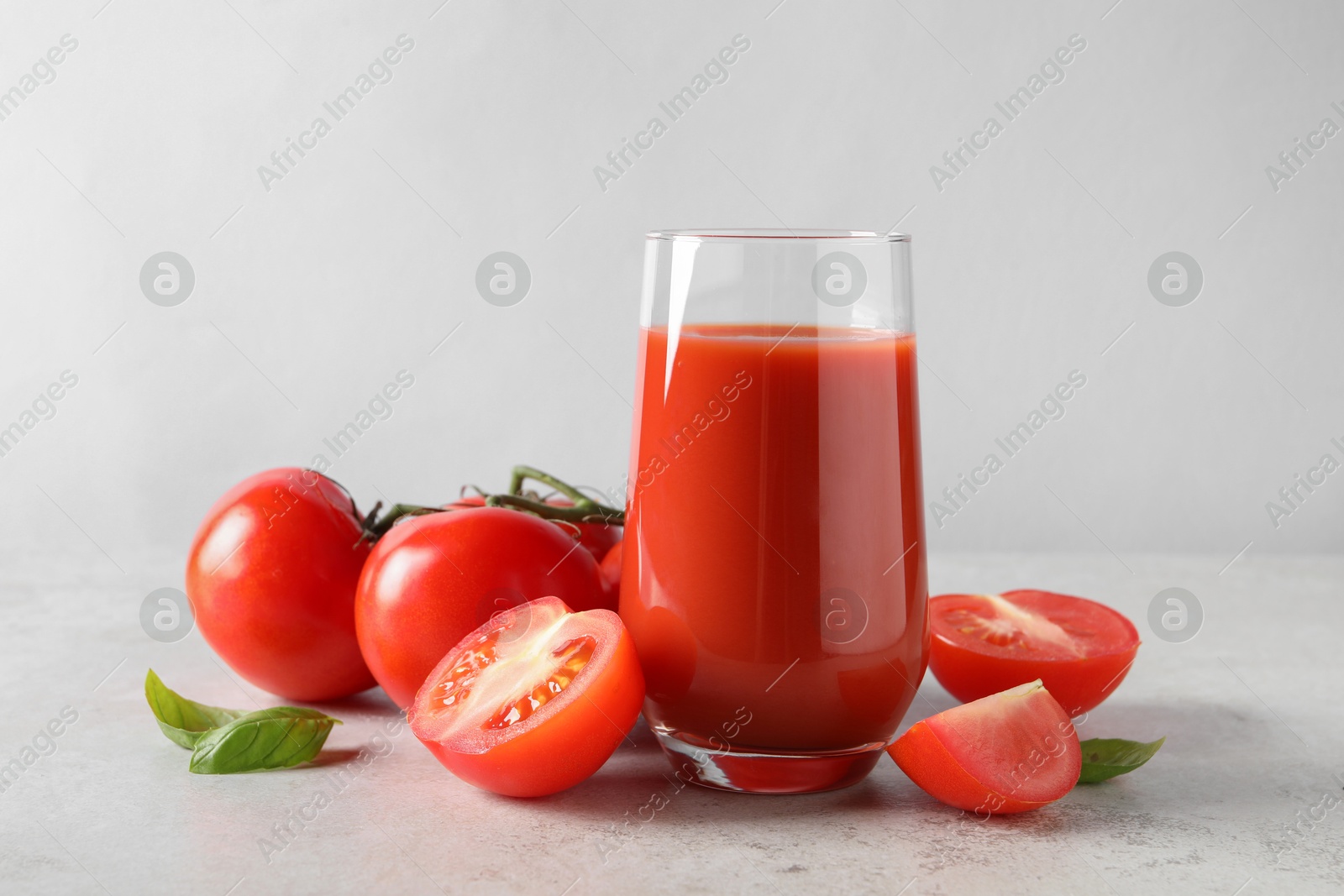 Photo of Tasty tomato juice in glass, basil leaves and fresh vegetables on light grey table