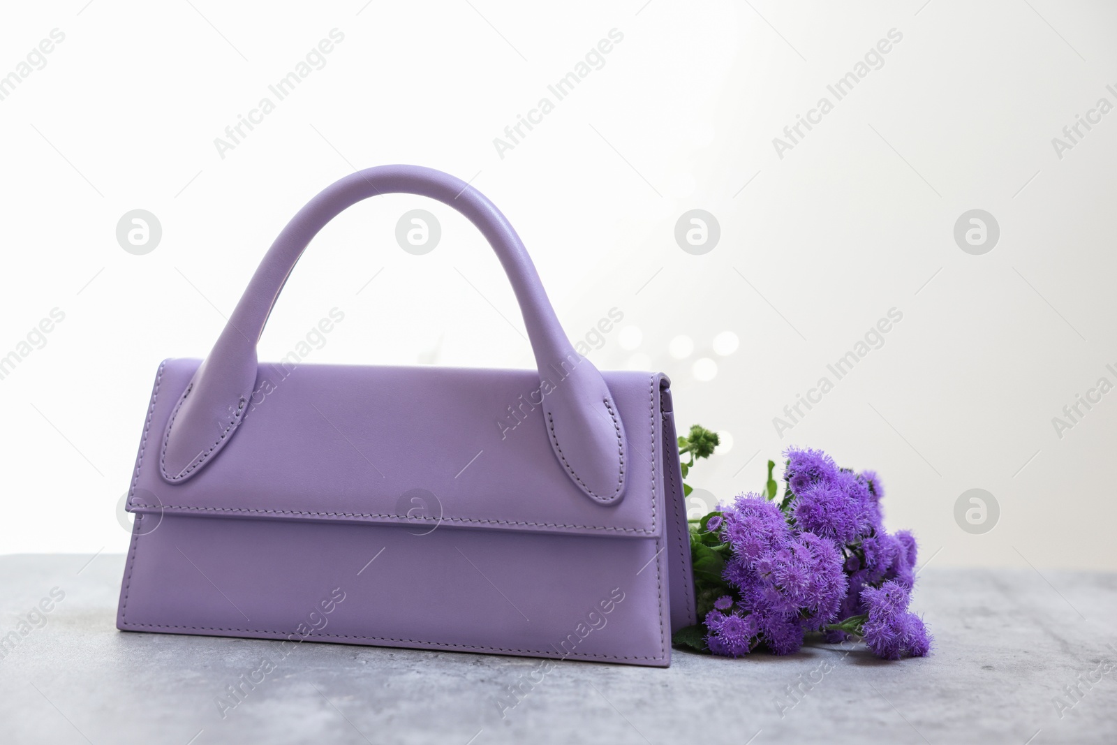 Photo of Stylish violet bag and ageratum flowers on gray table against light background, space for text