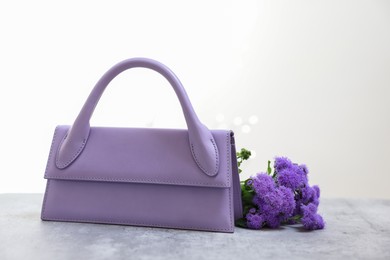 Stylish violet bag and ageratum flowers on gray table against light background, space for text