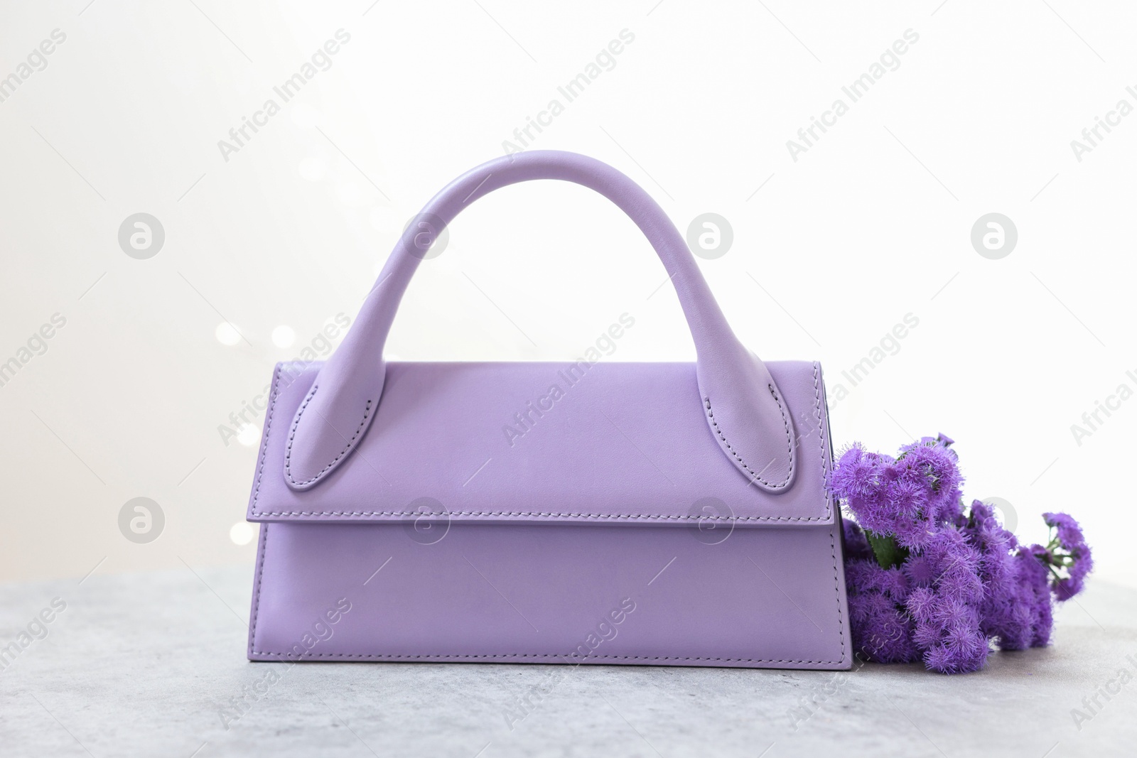 Photo of Stylish violet bag and ageratum flowers on gray table against light background