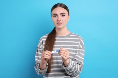 Sad woman holding pregnancy test on light blue background