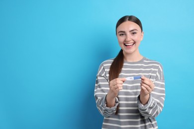 Happy woman holding pregnancy test on light blue background, space for text