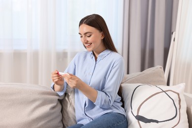 Happy woman holding pregnancy test on sofa indoors