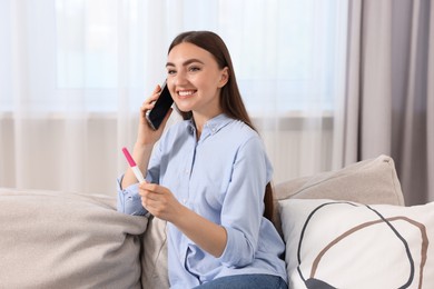 Happy woman with pregnancy test talking on smartphone indoors