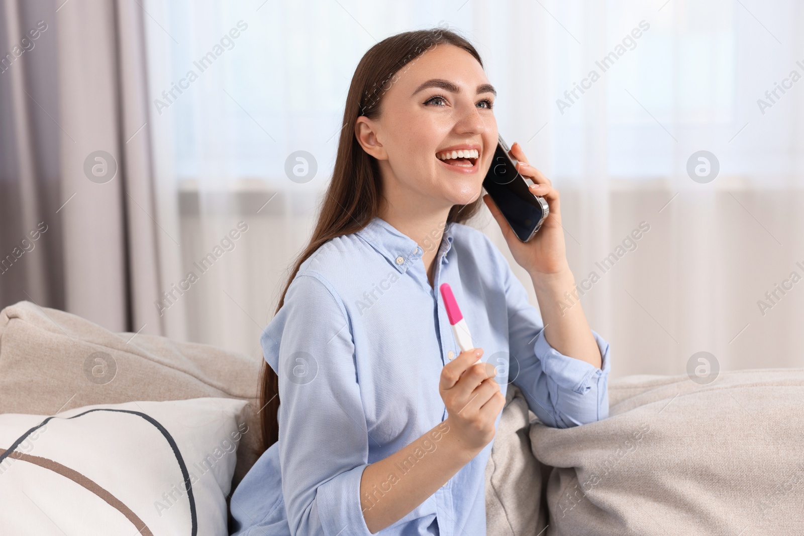 Photo of Happy woman with pregnancy test talking on smartphone indoors