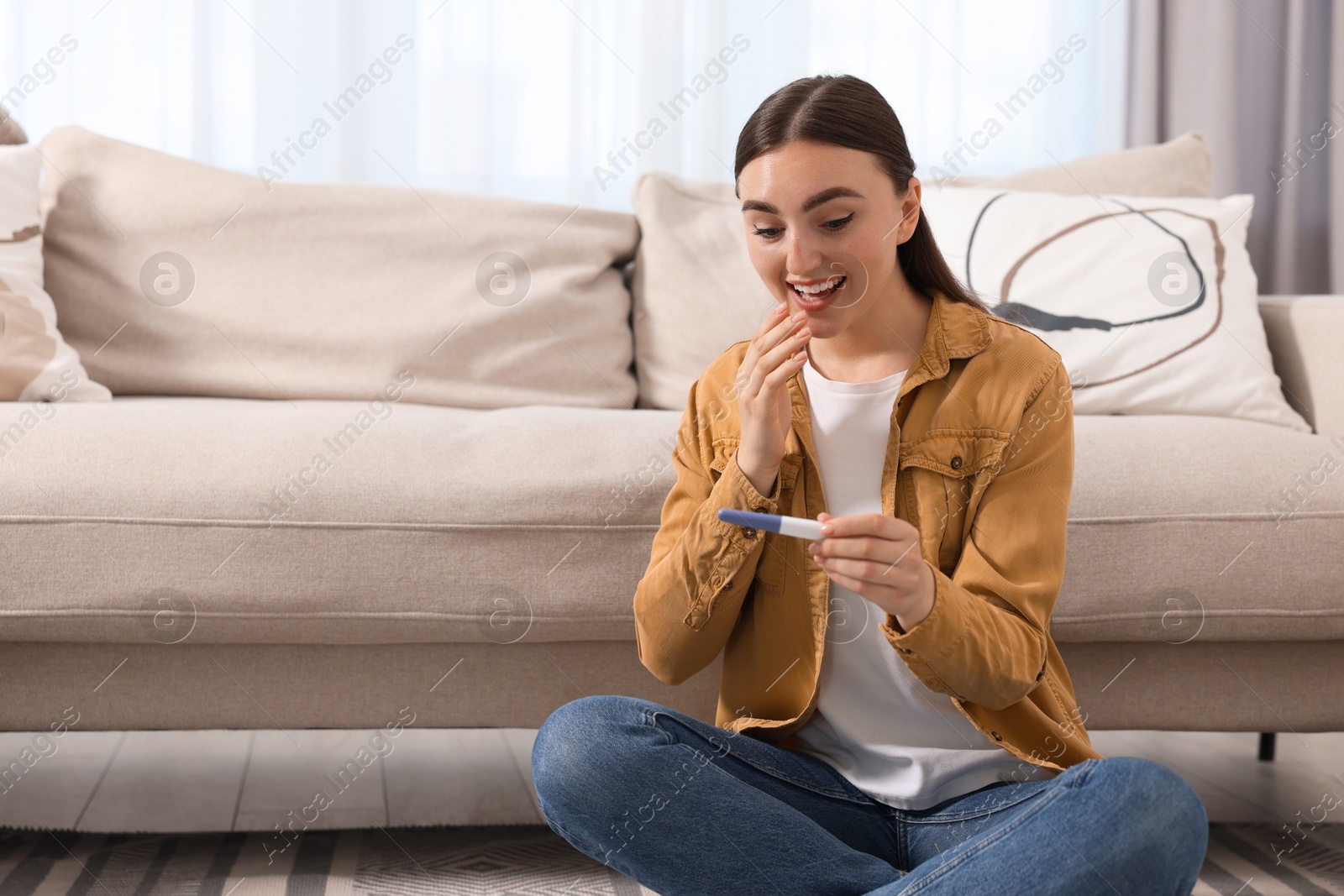 Photo of Happy woman holding pregnancy test on floor indoors