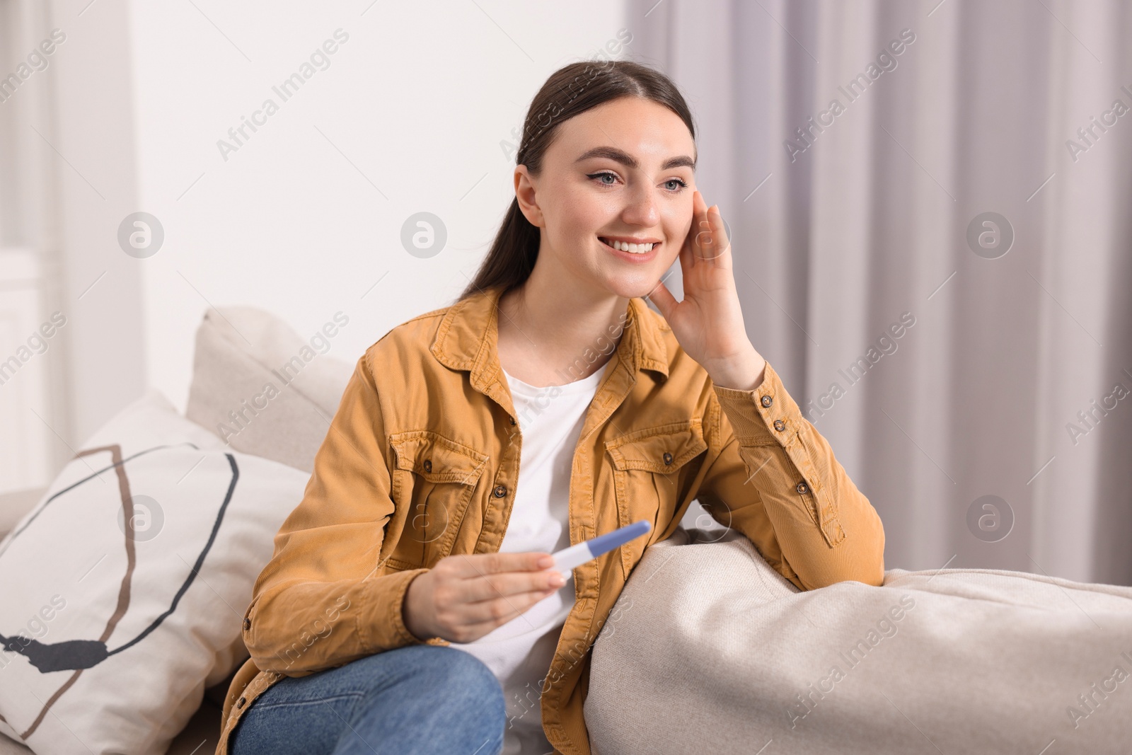 Photo of Happy woman holding pregnancy test on sofa indoors