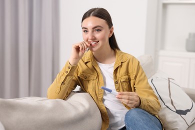 Happy woman holding pregnancy test on sofa indoors