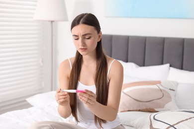 Sad woman holding pregnancy test on bed in room