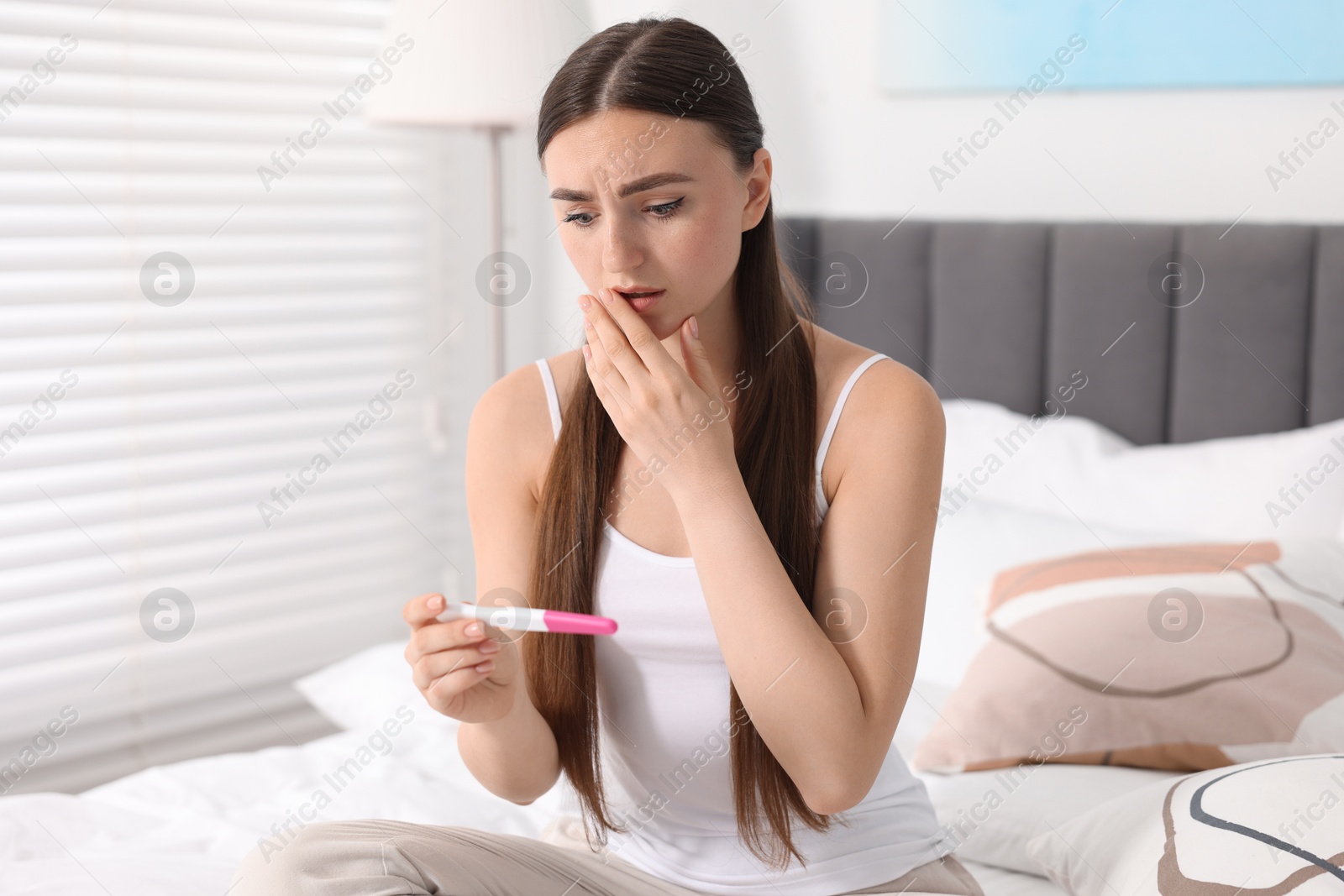 Photo of Shocked woman holding pregnancy test on bed in room