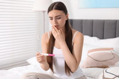 Shocked woman holding pregnancy test on bed in room