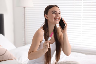 Happy woman with pregnancy test talking on smartphone in room